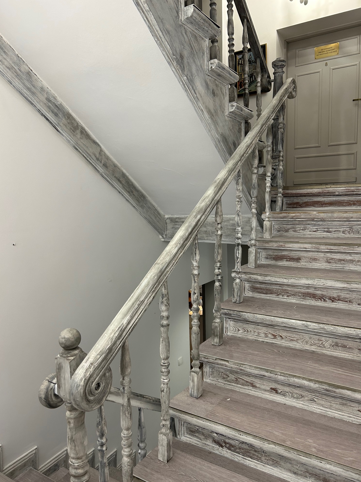 Drewniane schody z szarą, postarzaną balustradą prowadzące do szarych drzwi z numerem 8 i tabliczką informacyjną, widoczne detale wykończenia i efekt patyny.