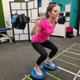 Kobieta w różowej koszulce wykonuje przysiad z obciążeniem na barkach, stojąc na dwóch niebieskich poduszkach równoważnych w siłowni.