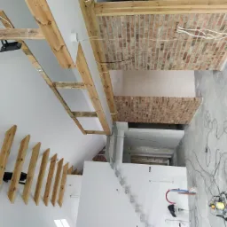 Wnętrze w trakcie prac wykończeniowych: widoczne drewniane elementy konstrukcyjne sufitu, fragment ściany wyłożonej cegłą, białe ściany, kabel elektryczny zwisający z sufitu i sprzęt budowlany...