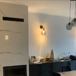 Salon z obudową kominka z płyt imitujących marmur, czarnym wkładem kominkowym, czarną szafką pod ścianą, dekoracjami i lampami wiszącymi.