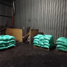 Stosy worków z opałem na paletach w magazynie z blachy falistej, widoczne opony i drewniane płyty w tle.