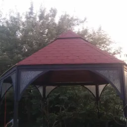 Altana ogrodowa z czerwoną dachówką bitumiczną na tle drzew, widoczne szklane panele między filarami, oświetlona zachodzącym słońcem.