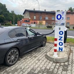 O-ZONIC sp. z o.o - Ciemnoszary samochód osobowy podłączony wężem do stacji O3 do ozonowania wnętrza pojazdu na zewnątrz, na brukowanym placu, w tle budynki mieszkalne.