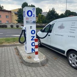 O-ZONIC sp. z o.o - Stanowisko do ozonowania pojazdów z podłączonym wężem do wnętrza białego samochodu dostawczego z logo firmy Revolution Technology na tle ulicy.