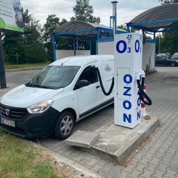O-ZONIC sp. z o.o - Biały van Dacia podłączony wężem do stacji ozonowania z napisem OZON. Stacja znajduje się na zewnątrz, na kostce brukowej. W tle widoczne zadaszone stanowiska.