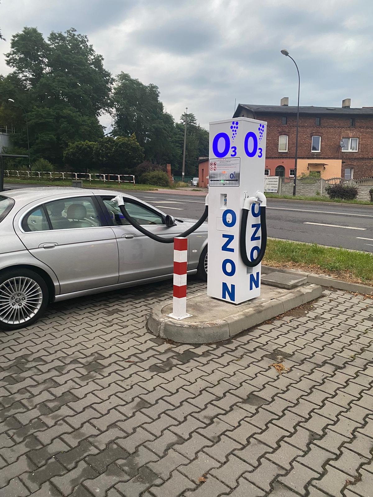 Samochód osobowy podłączony do stacji ozonowania na zewnątrz, widoczne przewody w oknach pojazdu, stacja z napisem OZON i symbolami O3, obok droga i budynek.
