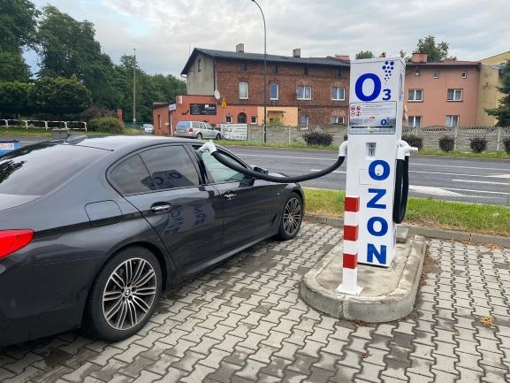 Ciemnoszary samochód osobowy podłączony wężem do stacji O3 do ozonowania wnętrza pojazdu na zewnątrz, na brukowanym placu, w tle budynki mieszkalne.