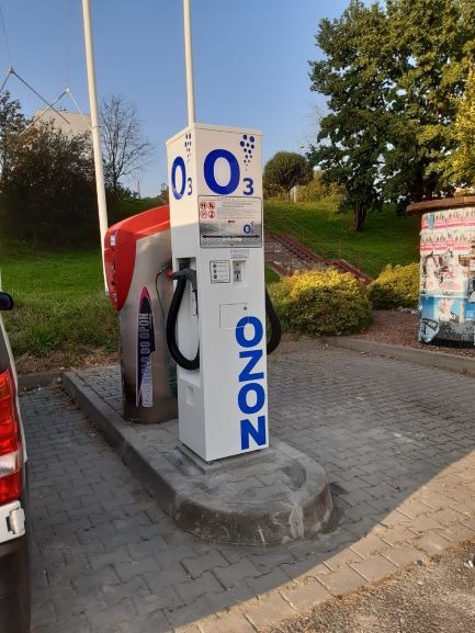 Ozonator samochodowy na zewnątrz, widoczny automat z napisem 'OZON' i symbolem O3, obok odkurzacz samochodowy, na kostce brukowej.
