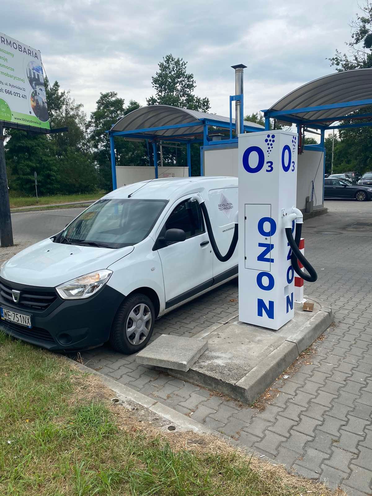 Biały van Dacia podłączony wężem do stacji ozonowania z napisem OZON. Stacja znajduje się na zewnątrz, na kostce brukowej. W tle widoczne zadaszone stanowiska.