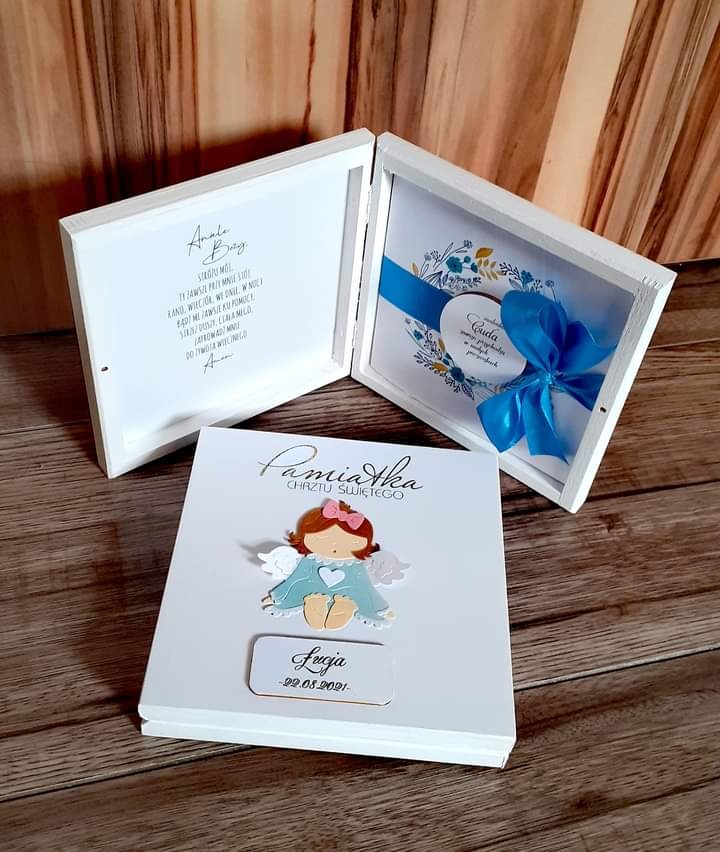 Ręcznie robiona pamiątka Chrztu Świętego w białym pudełku z aniołkiem, imieniem dziecka i datą, obok otwarte pudełka z dedykacją i kartką z niebieską wstążką.