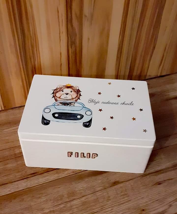 Białe, drewniane pudełko z imieniem 'FILIP', ozdobione rysunkiem lwa w samochodzie i złotymi gwiazdkami, na drewnianym tle.