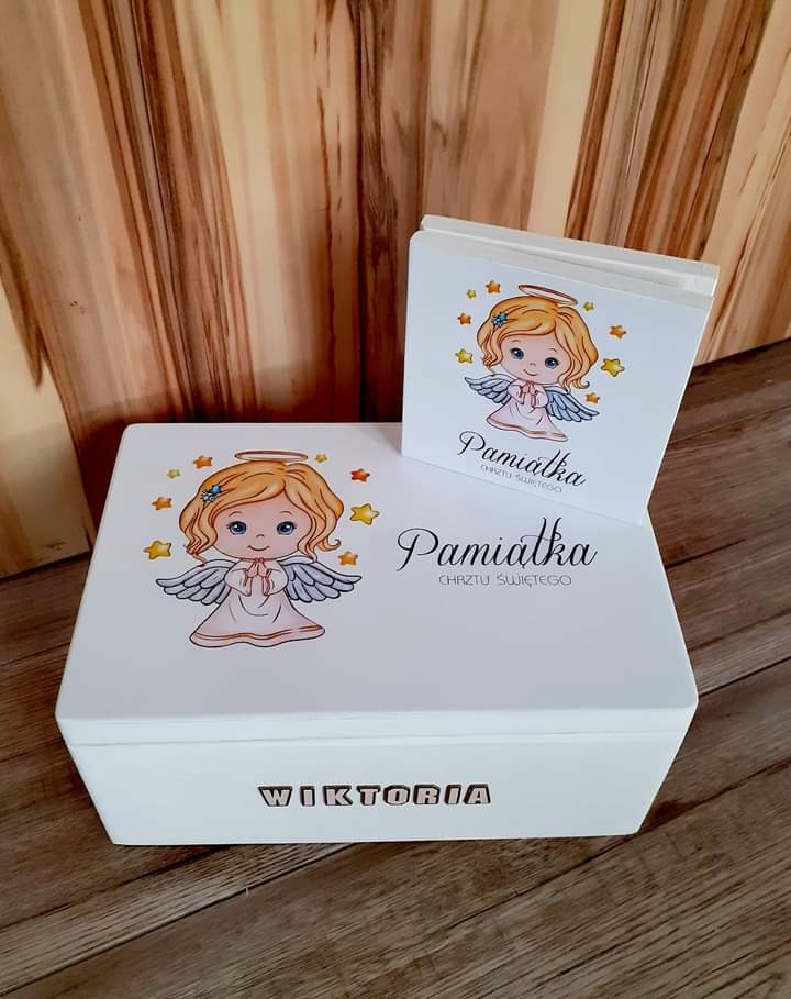 Białe pudełko i album pamiątkowy z motywem aniołka i napisem 'Pamiątka Chrztu Świętego' oraz imieniem 'Wiktoria', ustawione na drewnianej podłodze.
