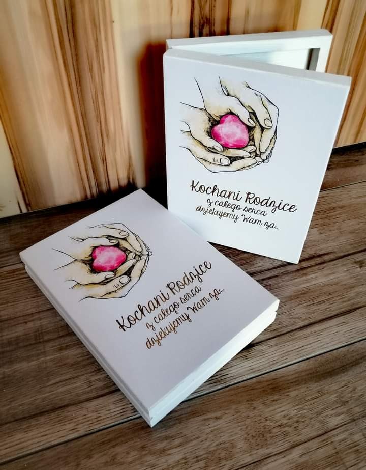 Dwa białe, drewniane pudełka z grafiką dłoni trzymających różowe serce i dedykacją dla rodziców, leżące na drewnianym stole.