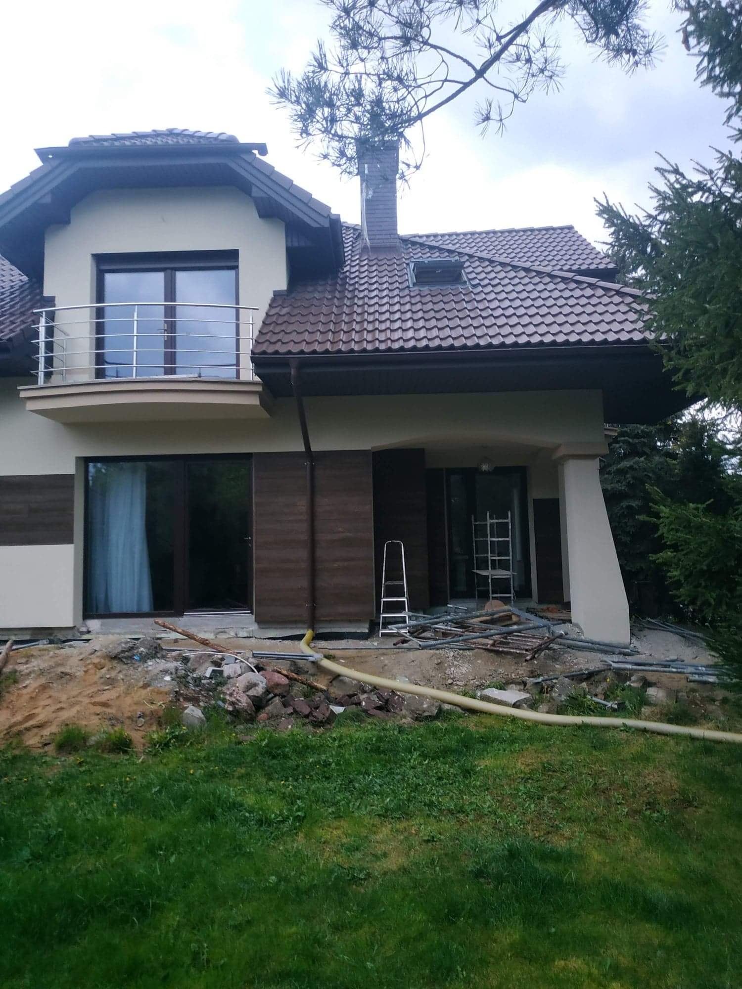 Dwukondygnacyjny dom z brązowym dachem w trakcie prac wykończeniowych elewacji, z widocznymi rusztowaniami i materiałami budowlanymi wokół budynku.