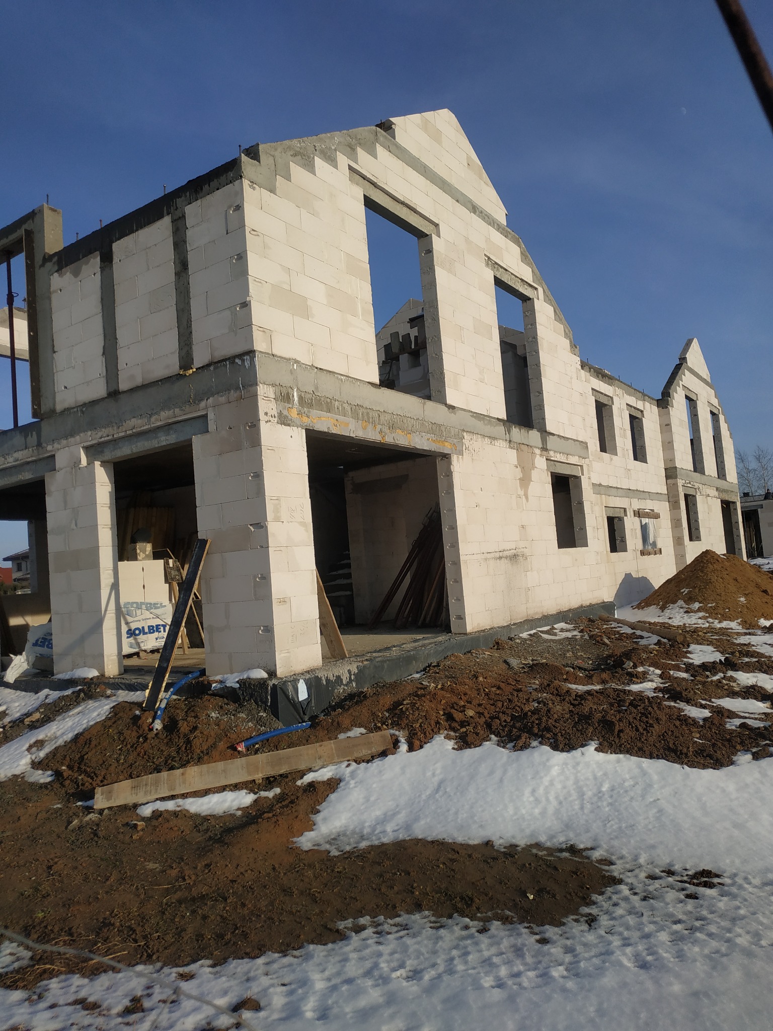 Surowy stan domu jednorodzinnego w trakcie budowy z białych bloczków, widoczne otwory okienne i drzwiowe, na ziemi resztki śniegu i sterta ziemi.