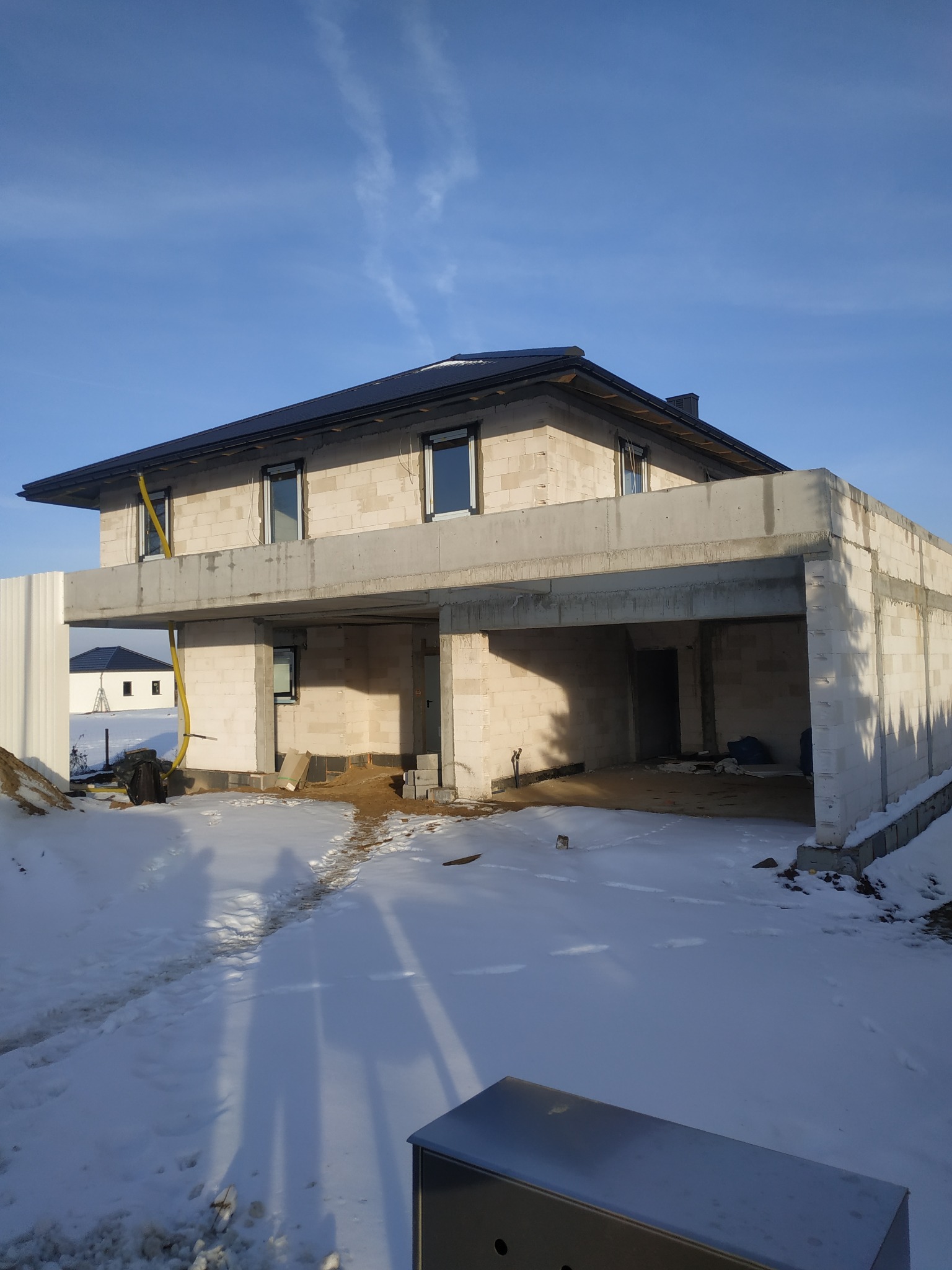 Surowy stan domu w trakcie budowy, ściany z betonu komórkowego, widoczne okna i zadaszenie nad wjazdem do garażu, teren wokół zasypany śniegiem, żółty wąż budowlany zwisający z okna.