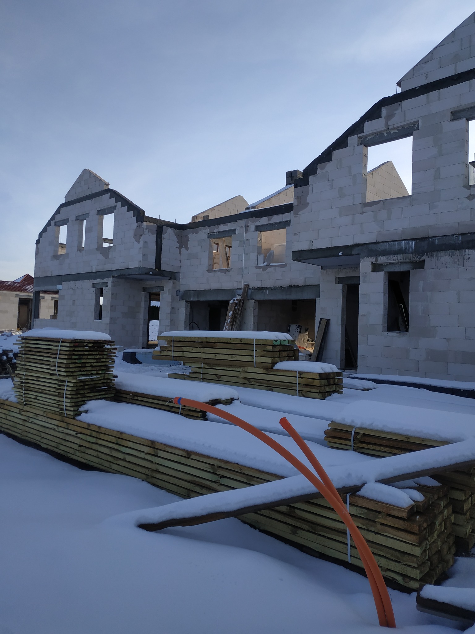 Surowe budynki w stanie surowym zamkniętym z bloczków, drewno budowlane na pierwszym planie pokryte śniegiem, pomarańczowe rury wystają z zaspy.
