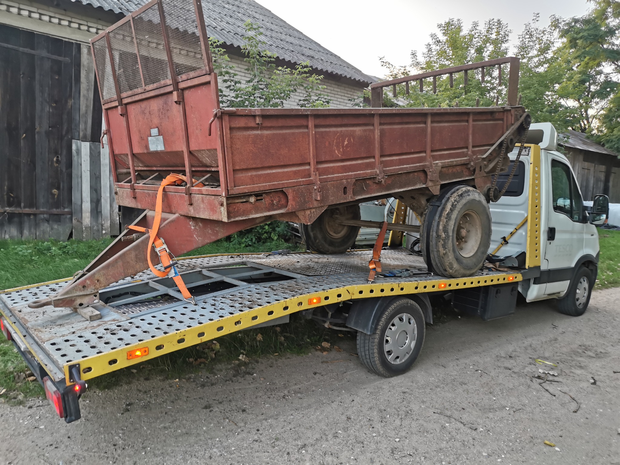 Biała ciężarówka z żółtą platformą przewozi rdzewiejącą, czerwoną przyczepę rolniczą zabezpieczoną pomarańczowymi pasami.