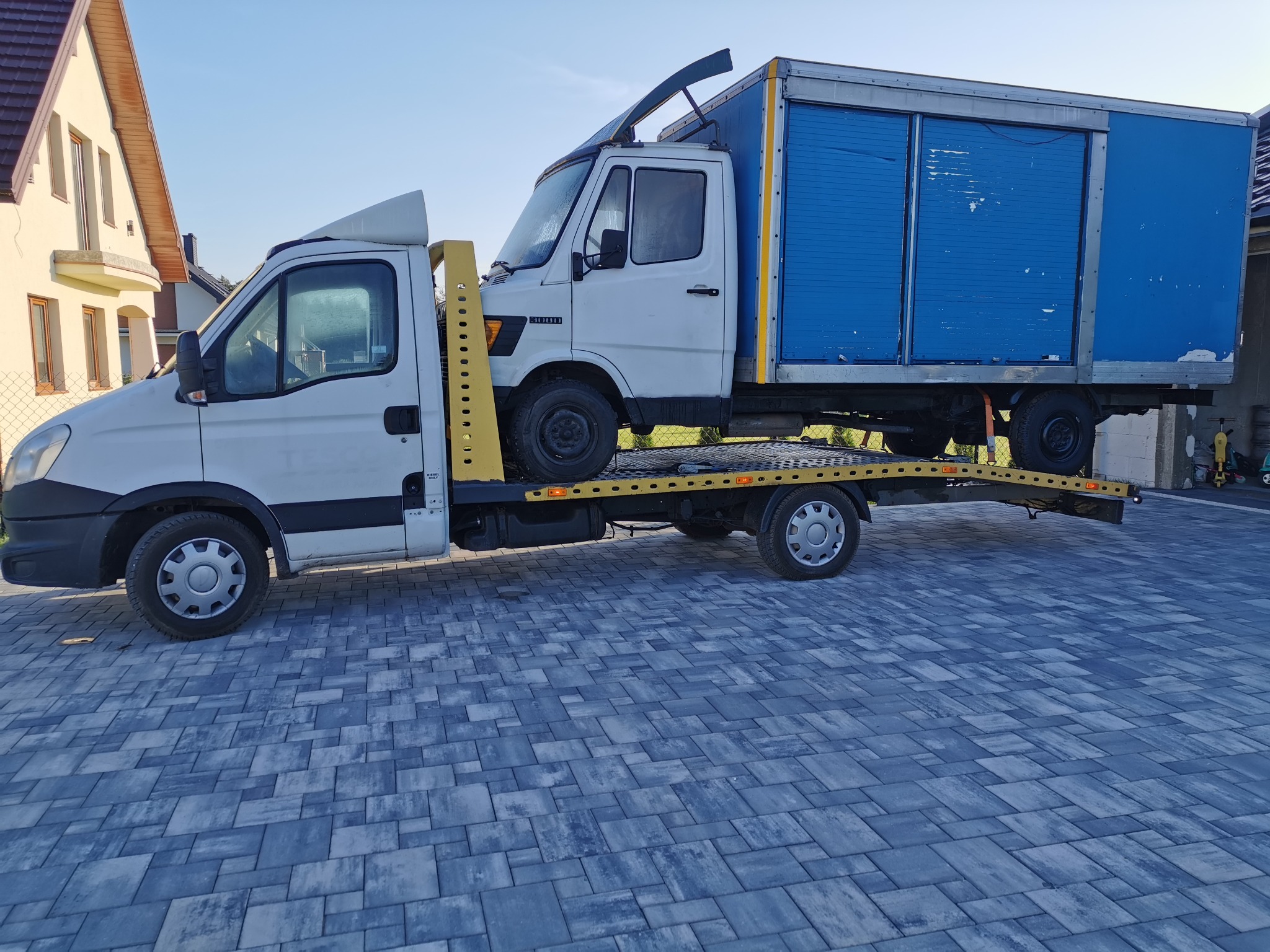 Biała platforma pomocy drogowej przewozi białego dostawczaka z niebieską zabudową kontenerową na tle kostki brukowej i budynku.