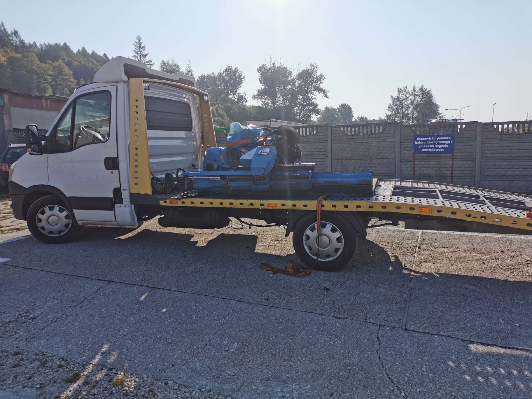 Biała ciężarówka z żółtą konstrukcją lawety przewozi niebieską maszynę przemysłową zabezpieczoną pasami na tle betonowego ogrodzenia i drzew.