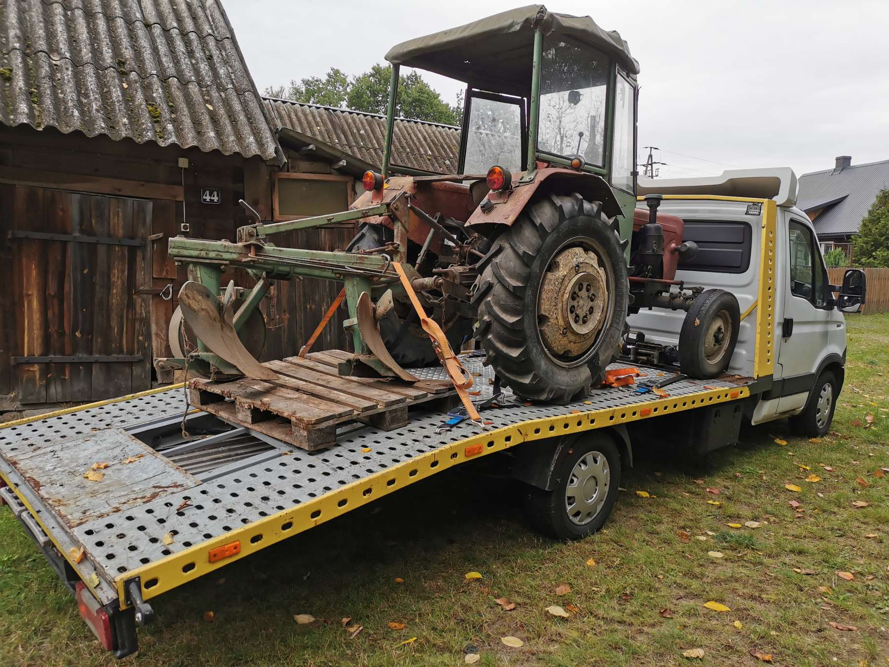 Stary traktor z pługiem zabezpieczony pasami na platformie lawety, na tle drewnianego budynku.