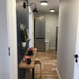 Dream house group Sp. z o.o. - Korytarz po remoncie z drewnopodobną podłogą, granatową ścianą z półką, na której stoją filiżanki i dekoracje, oraz widoczną kuchnią w tle.