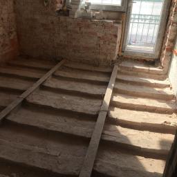 Dream house group Sp. z o.o. - Surowe, ceglane wnętrze w trakcie przebudowy z widocznymi schodami w stanie surowym i oknem z widokiem na zewnątrz, cegły ułożone na parapecie okiennym.