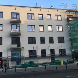 Dream house group Sp. z o.o. - Budynek wielorodzinny w trakcie prac elewacyjnych, widoczne rusztowania, siatka ochronna, pracownicy oraz częściowo odnowiona fasada z nowymi oknami.