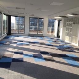 Dream house group Sp. z o.o. - Jasne biuro z nową, wzorzystą wykładziną dywanową w odcieniach szarości i błękitu, duże okna z widokiem na budynek biurowy, wpadające promienie słoneczne.