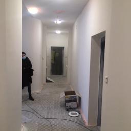 Dream house group Sp. z o.o. - Długi korytarz w trakcie malowania na biało, widoczna osoba w maseczce i karton po urządzeniu, podłoga zabezpieczona folią.