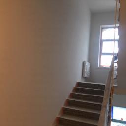 Dream house group Sp. z o.o. - Klatka schodowa w trakcie malowania: zabezpieczone schody, świeżo pomalowane ściany w jasnym odcieniu, okno z widocznym rusztowaniem na zewnątrz.