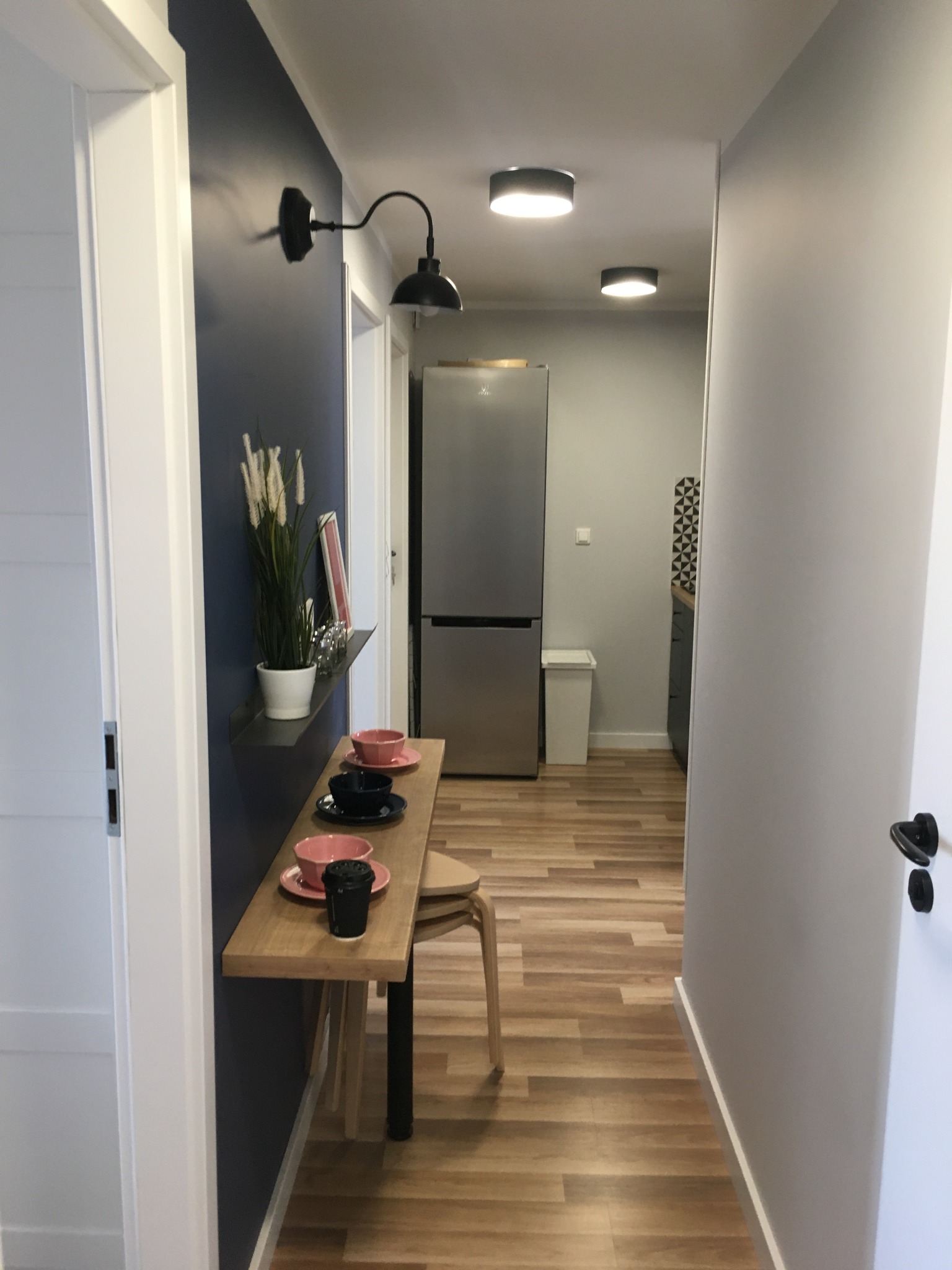 Korytarz po remoncie z drewnopodobną podłogą, granatową ścianą z półką, na której stoją filiżanki i dekoracje, oraz widoczną kuchnią w tle.