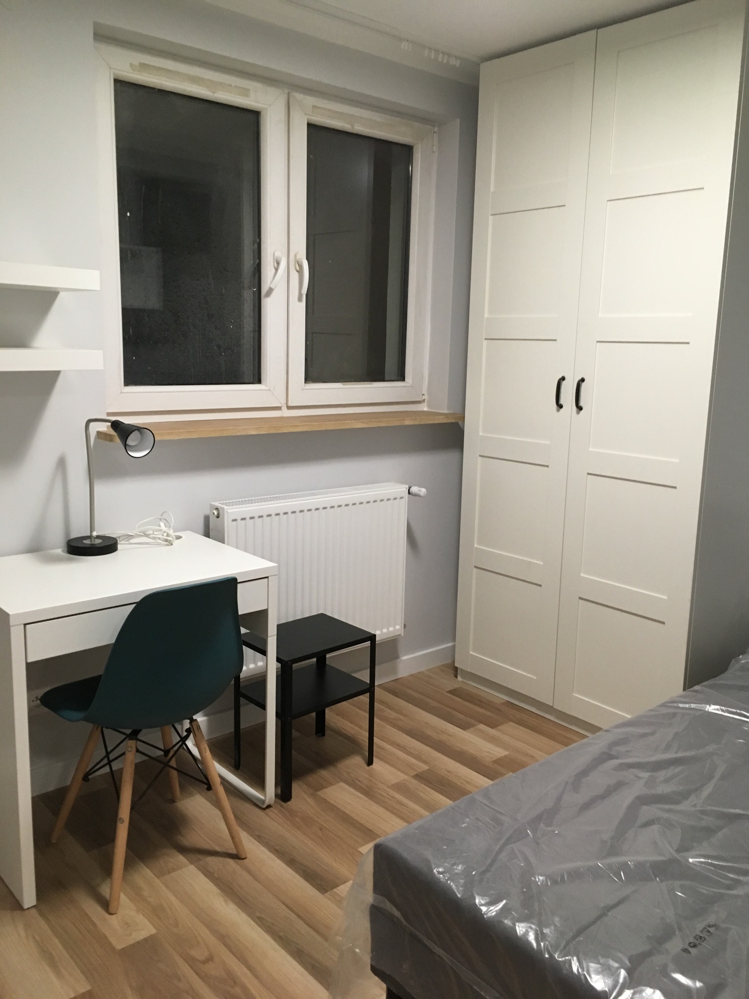 Pokój po odświeżeniu: białe ściany, jasna podłoga, nowe biurko z lampką, krzesło, szafa i łóżko przykryte folią ochronną.