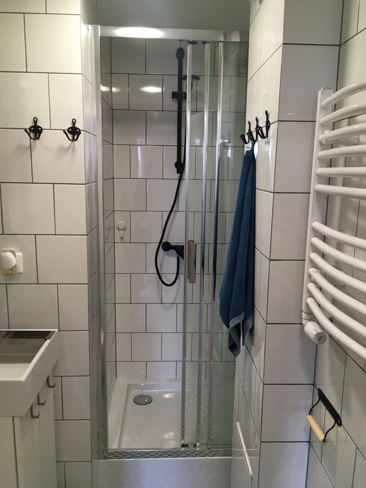 Biała łazienka z kabiną prysznicową typu walk-in, czarną armaturą i grzejnikiem drabinkowym; widoczny ręcznik w odcieniach niebieskiego.