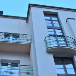 Odnowiona elewacja budynku wielorodzinnego z balkonami o zielonych barierkach, widok z dołu, obok budynku z nieodnowioną elewacją.