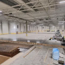 Rozległa hala przemysłowa w trakcie wylewania posadzki, widoczne zbrojenia, szalunki oraz maszyny budowlane w oddali.