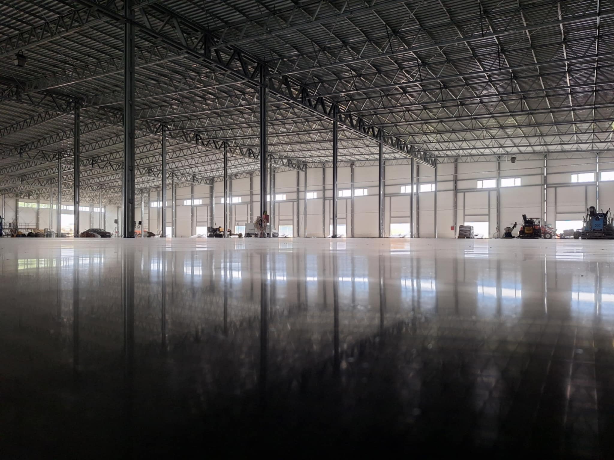 Błyszcząca, świeżo wylana posadzka w hali przemysłowej z widocznymi maszynami budowlanymi i refleksami świetlnymi na powierzchni.