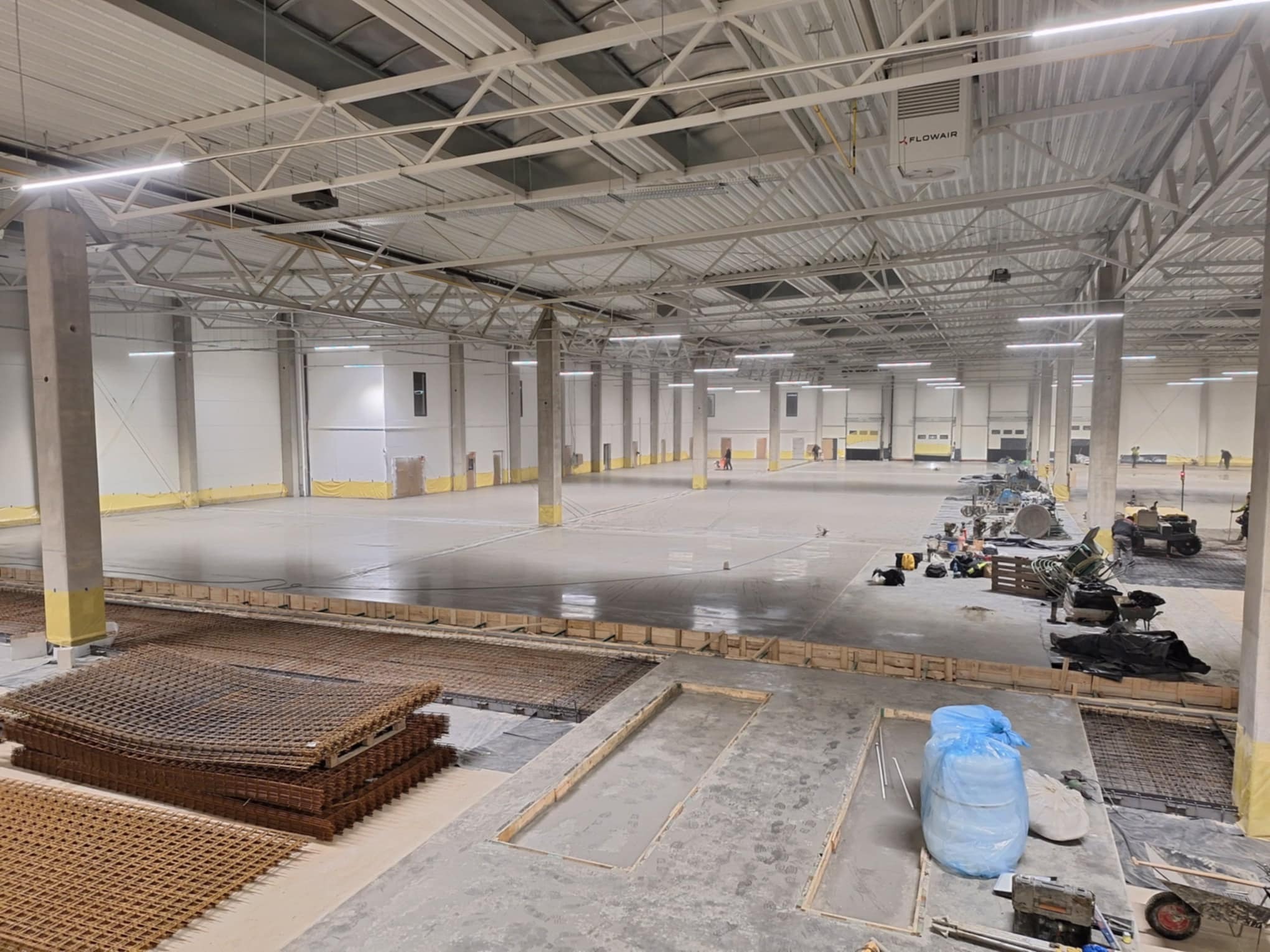 Rozległa hala przemysłowa w trakcie wylewania posadzki, widoczne zbrojenia, szalunki oraz maszyny budowlane w oddali.