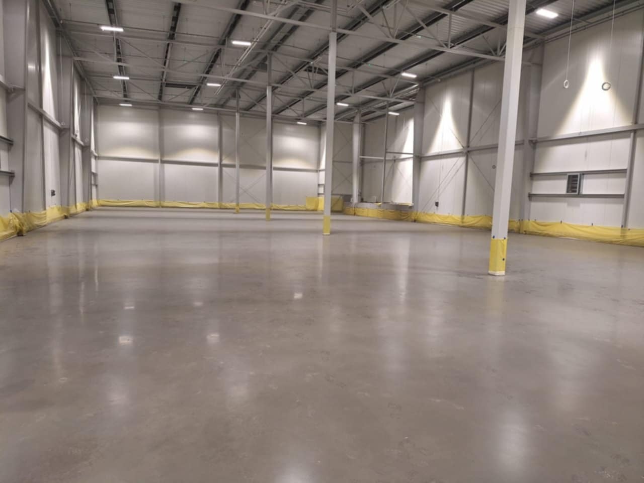 Wnętrze hali magazynowej z gładką, szarą posadzką, zabezpieczonymi żółtą folią słupami i ścianami, oświetlone górnymi lampami.