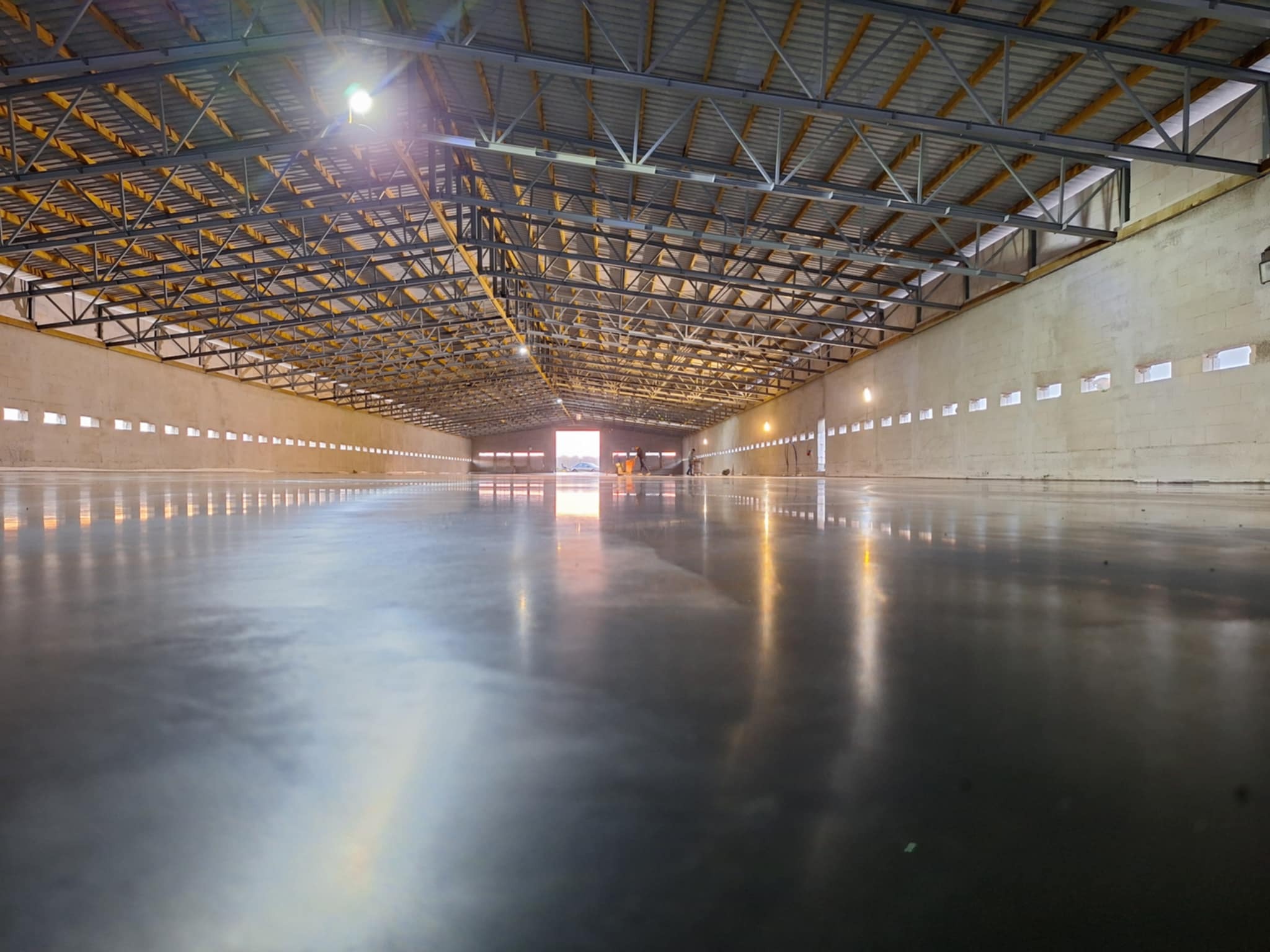 Wnętrze hali produkcyjnej z nową, błyszczącą posadzką, widoczne stalowe elementy konstrukcji dachu i rzędy okien w ścianach.