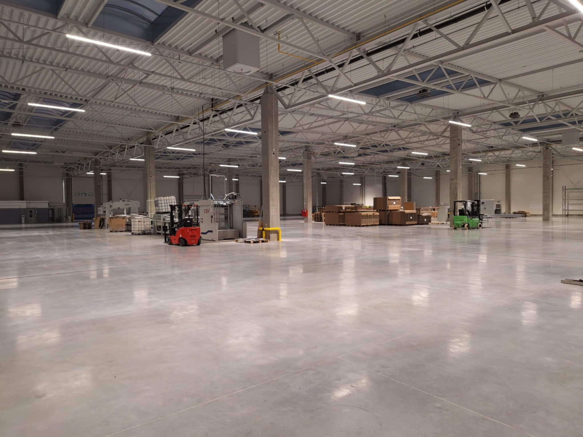 Rozległa hala magazynowa z nową, gładką, szarą posadzką, widoczne wózki widłowe i składowane towary, oświetlenie sufitowe.