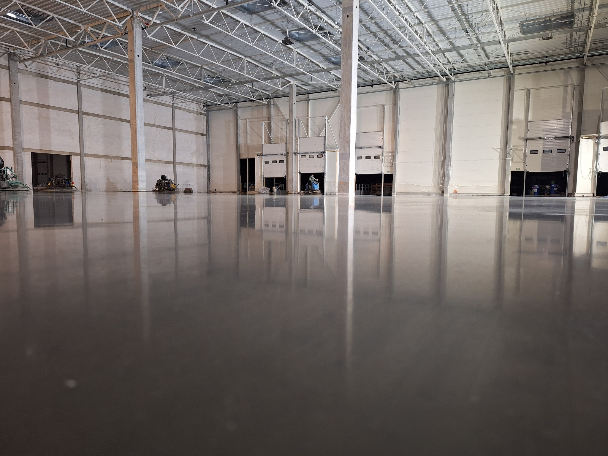 Błyszcząca, szara posadzka w hali przemysłowej z widocznymi maszynami do polerowania w tle i metalową konstrukcją dachu.