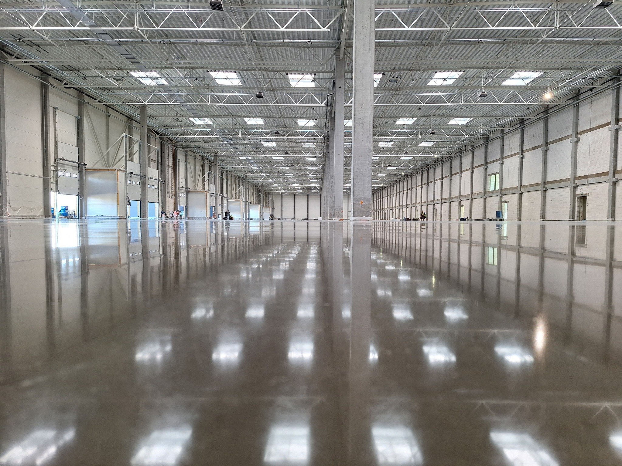 Błyszcząca, szara posadzka w hali magazynowej, odbijająca światło z okien dachowych i lamp, z widocznymi słupami konstrukcyjnymi i bramami w tle.