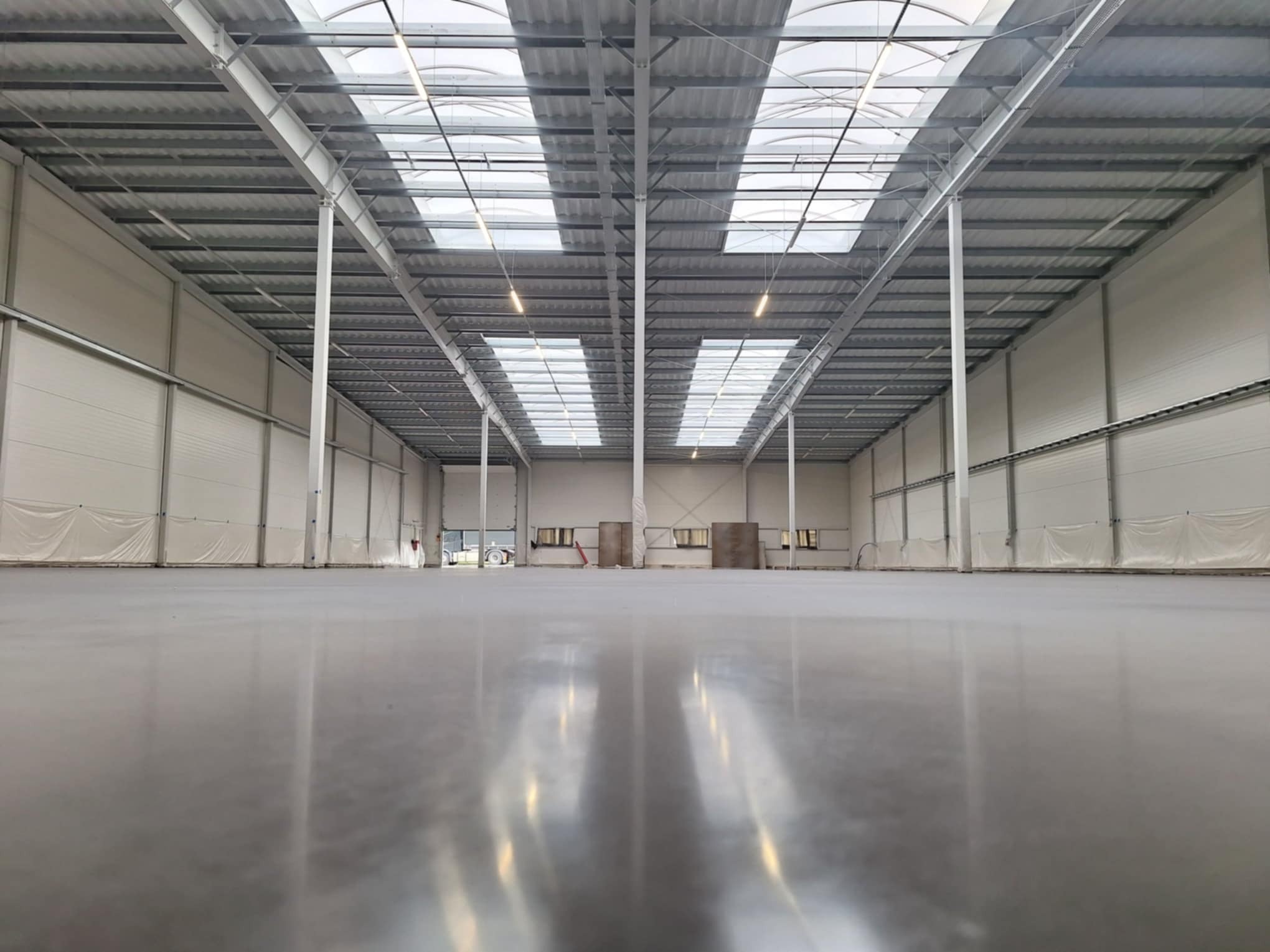 Błyszcząca, szara posadzka w nowej hali magazynowej z widoczną konstrukcją stalową dachu i świetlikami, oraz białymi ścianami z widocznymi zasłonami ochronnymi.