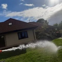Strumień wody z węża podlewa trawnik przed domem z czerwoną dachówką, widoczne niebieskie niebo z chmurami i słońce w kadrze.