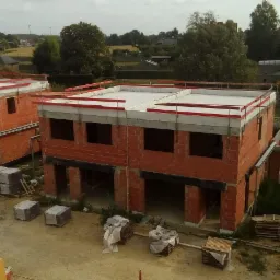 Wznoszenie ścian dwukondygnacyjnych domów z czerwonej cegły na osiedlu, widok z góry, materiały budowlane na paletach w pobliżu.