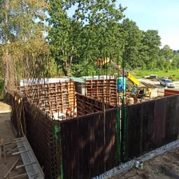 Budowa fundamentów domu: szalunek zbrojony stalą, pracownik w czerwonej koszulce wewnątrz konstrukcji, żółta koparka z podnośnikiem w tle, teren budowy z drzewami.