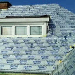 Montaż izolacji termicznej ISOTEC na dachu domu z oknem dachowym, widoczne fragmenty starego pokrycia dachowego z cegły oraz fragment nieba.