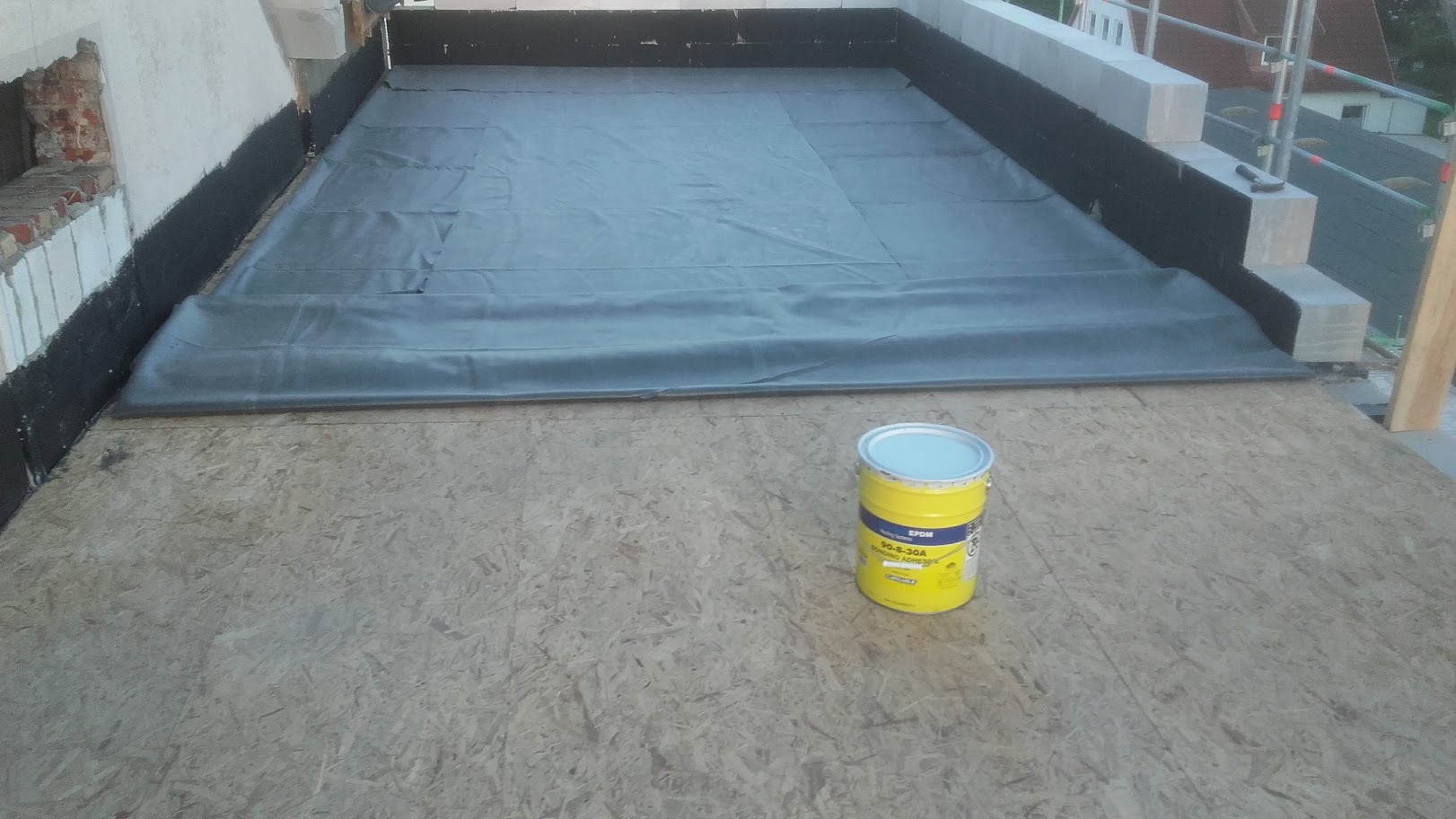 Płaska powierzchnia dachu pokryta szarą membraną EPDM, z widocznym żółtym pojemnikiem z klejem EPDM, obróbkami blacharskimi i ścianą w tle.