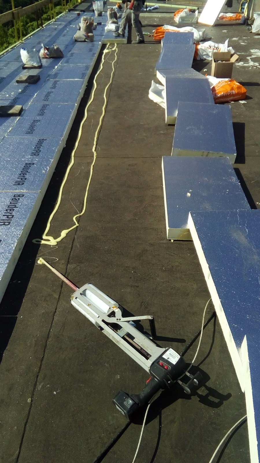 Montaż izolacji termicznej na dachu płaskim: płyty styropianowe pokryte folią aluminiową, aplikacja kleju montażowego za pomocą pistoletu akumulatorowego, widoczne worki z materiałami i pracownik...